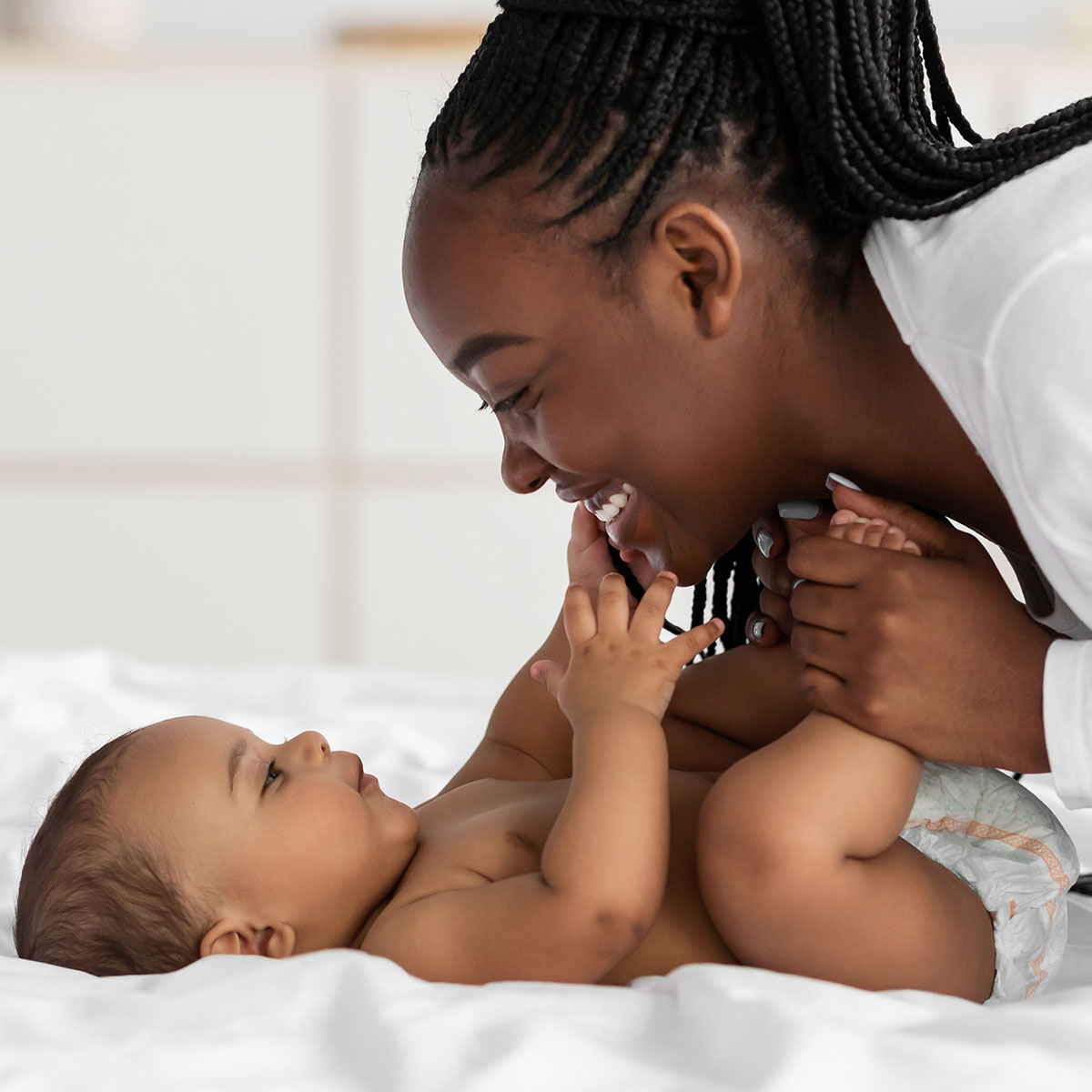 A woman is smiling at her baby who is lying on a bed. She has the baby's feet gently in her hands whilst the baby is touching her face.