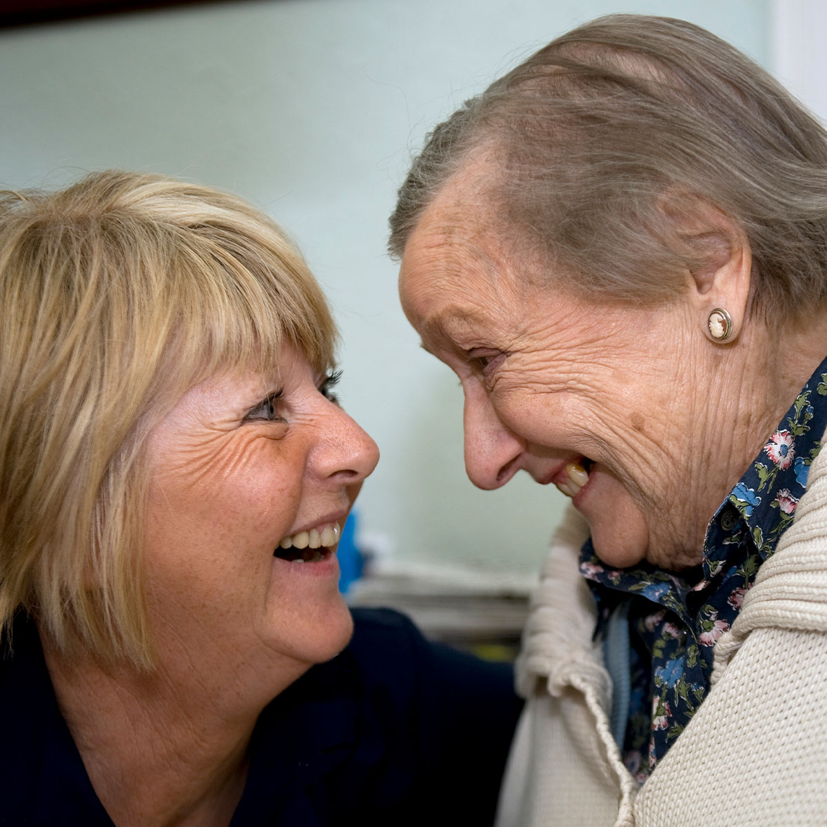 A female nurse is crouched down and smiling looking face to face with a female pensioner who is also smiling.