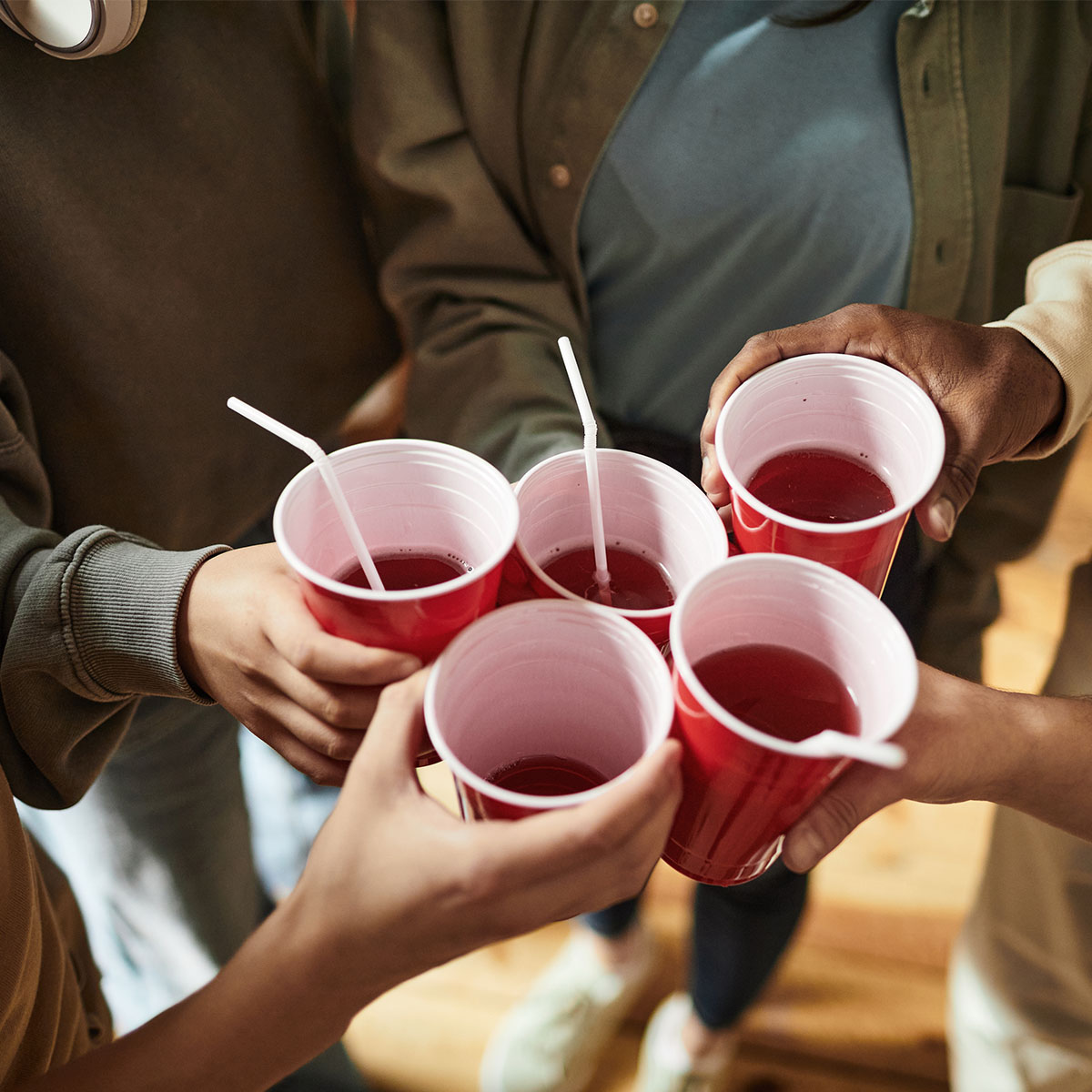 Five young people are giving a toast with plastic cups. Three have straws in whilst the other two cups do not. We cannot see the young people's faces, just their hands.