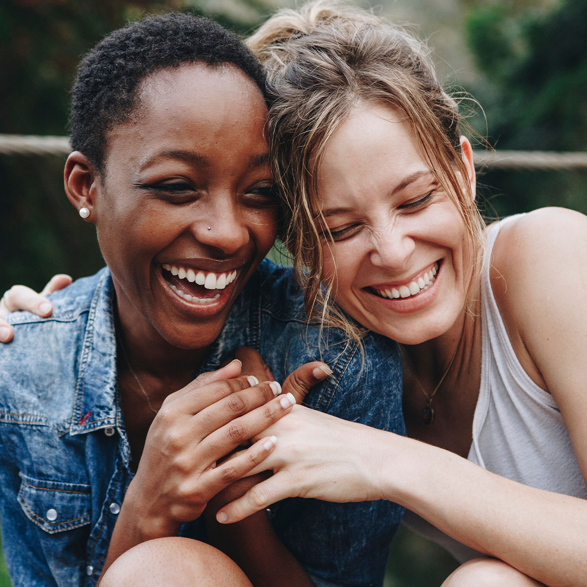 Two women in their twenties are laughing. The woman on the right has her arm around the one of the left. The woman on the left is holding the other woman's free hand. They are resting their heads on each others.